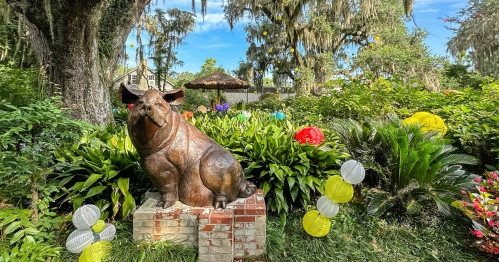 A bronze pig statue sits on a brick pedestal surrounded by colorful flowers and lanterns in a lush garden.