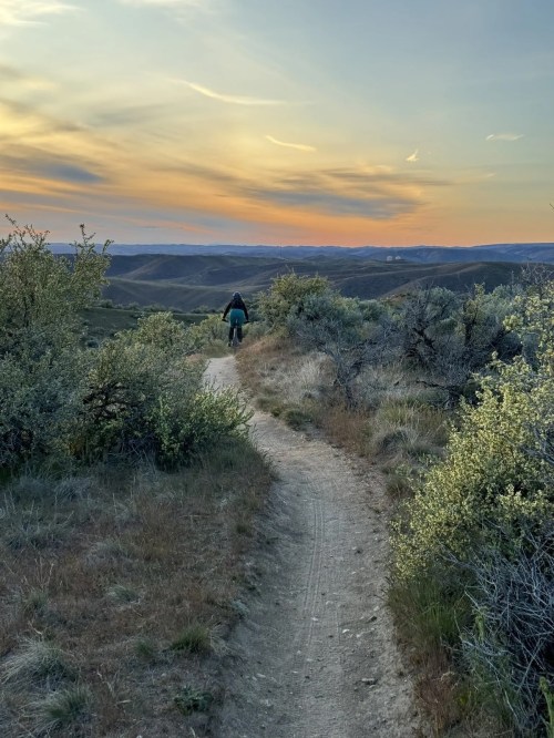 A person biking on a winding trail through grassy hills under a colorful sunset sky.