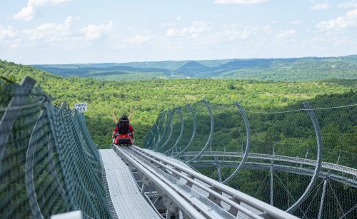 A person rides a mountain coaster on a track overlooking lush green hills and a blue sky with fluffy clouds.
