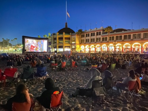 A crowd watches a movie on a large screen set up on the beach at night, with a historic building in the background.