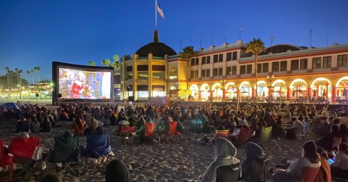 A large outdoor movie screen shows a film as a crowd sits on the beach, with colorful buildings and palm trees in the background.