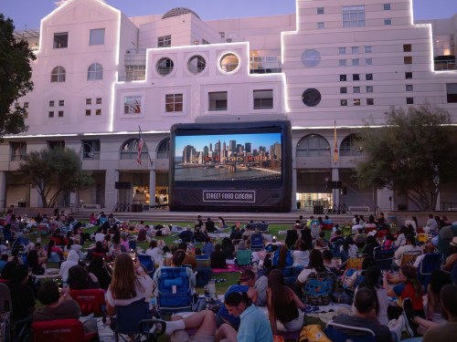 A large outdoor screen displays a city skyline, with a crowd seated on the grass enjoying a movie night.