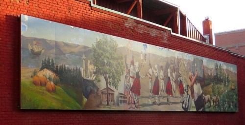 A colorful mural depicting historical scenes, including dancers, landscapes, and a ship, on a brick wall.
