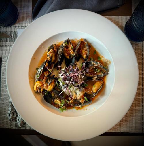 A plate of mussels in a savory sauce, garnished with microgreens, served on a white dish.