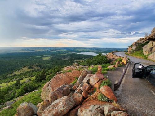 A scenic view from a rocky overlook, showcasing lush green hills and a lake under a cloudy sky.