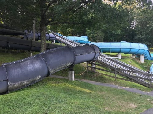 A winding water slide with black and blue sections, surrounded by green grass and trees in a park setting.