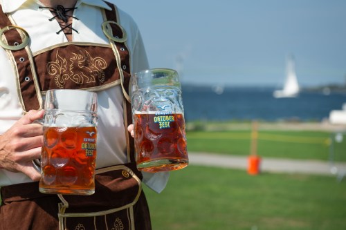 A person in traditional attire holds two large beer steins by a waterfront, with a sailboat in the background.