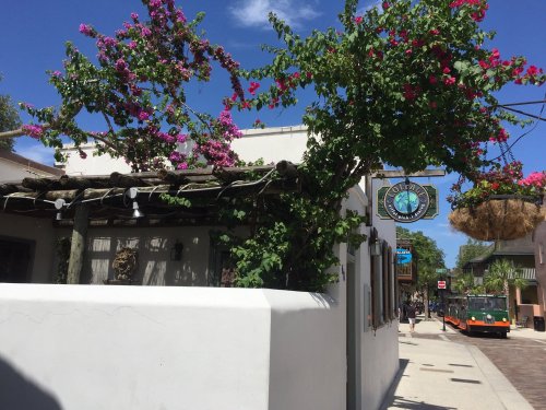 A sunny street scene featuring a white building adorned with vibrant flowers and a sign for a local business.