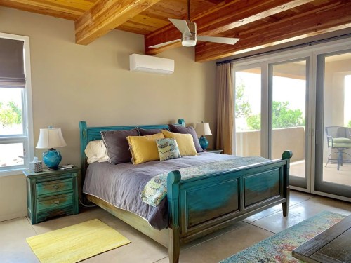 Bright bedroom with a teal bed, yellow pillows, wooden beams, and large windows leading to a balcony.