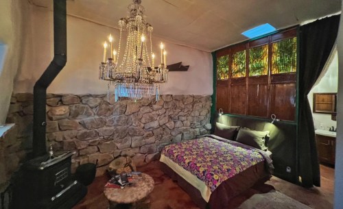 Cozy bedroom with a stone wall, chandelier, colorful bedding, and a small wood stove, creating a warm atmosphere.