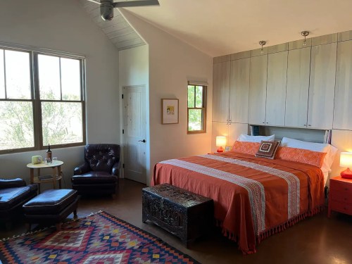 Cozy bedroom with a large bed, orange bedding, a leather chair, and a decorative rug, featuring natural light from windows.