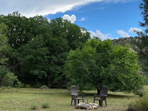 Two chairs and a small table near a fire pit, surrounded by lush greenery and a blue sky with clouds.