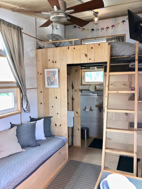 Cozy tiny home interior featuring a lofted bed, wooden accents, and a living area with a sofa and natural light.