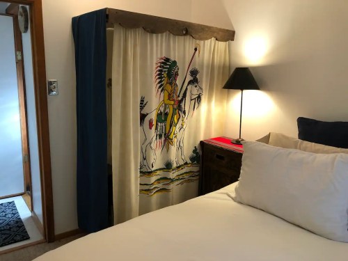 A cozy bedroom featuring a bed, a lamp, and a decorative curtain with a Native American motif.