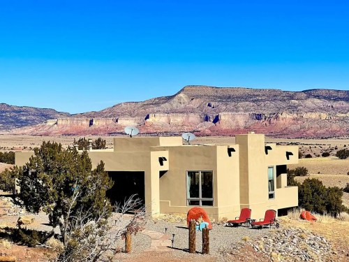 A modern adobe-style house with satellite dishes, surrounded by desert landscape and mountains under a clear blue sky.