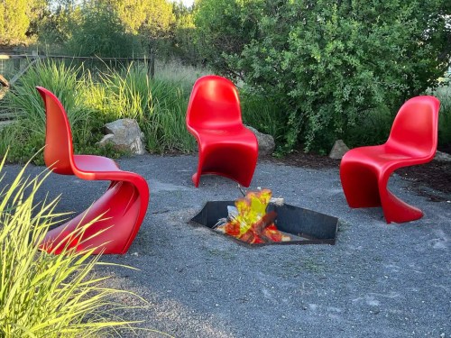 Four red chairs surround a fire pit in a gravel area, with greenery in the background.