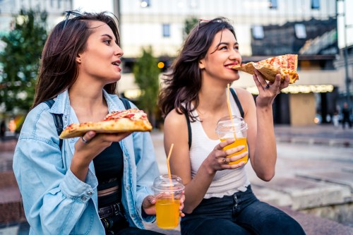 Two young women enjoy pizza and drinks while sitting outdoors, smiling and chatting in a lively urban setting.