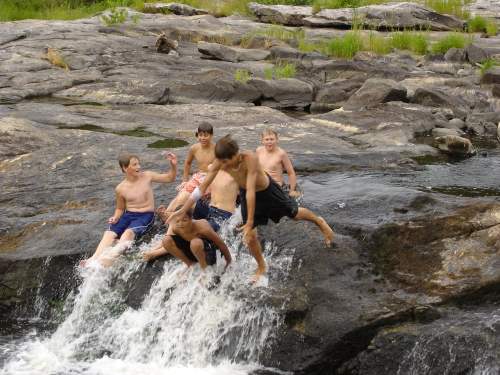 A group of boys playing on rocks by a waterfall, some jumping into the water while others sit nearby.