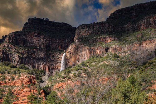 A waterfall cascades down rocky cliffs surrounded by lush greenery and dramatic clouds in a mountainous landscape.