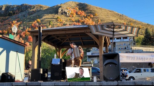 A musician plays accordion on stage under a gazebo, with autumn foliage on the hillside in the background.