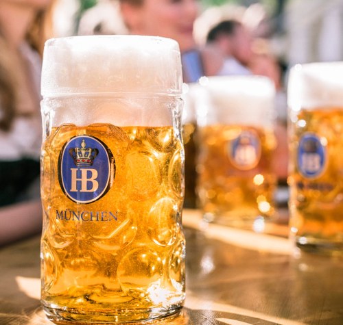 A close-up of a frothy beer mug with the HB München logo, surrounded by blurred glasses in the background.