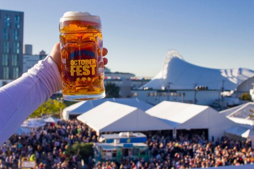 A hand holds a large beer mug at an outdoor festival, with a crowd and tents in the background.