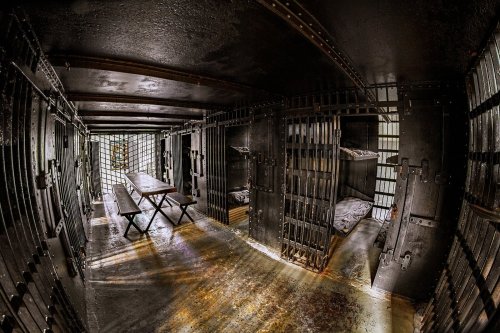 Interior of a dimly lit prison cell block with metal bars, a long table, and two empty bunks.