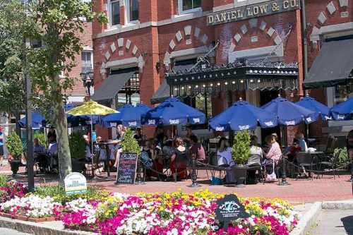 Outdoor café scene with patrons sitting under blue umbrellas, surrounded by colorful flowers and a historic brick building.