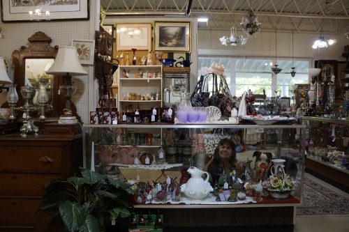 A vintage shop display featuring various antiques, lamps, and decorative items, with a person reflected in the glass.