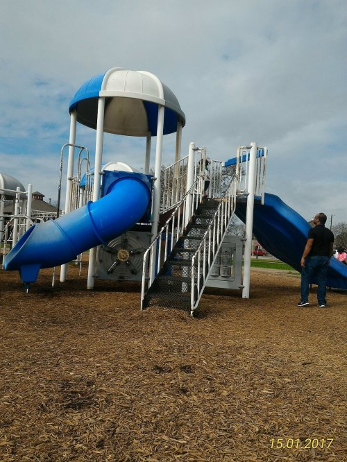 A playground featuring a blue slide, stairs, and a dome structure, with a person standing nearby on a wood chip surface.