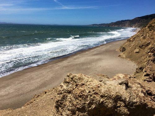 A scenic view of a sandy beach with waves crashing against the shore, framed by rocky cliffs under a clear blue sky.