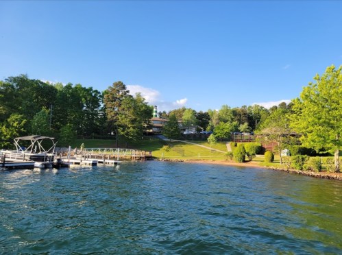 A serene lakeside view with a dock, green trees, and a house on a hill under a clear blue sky.