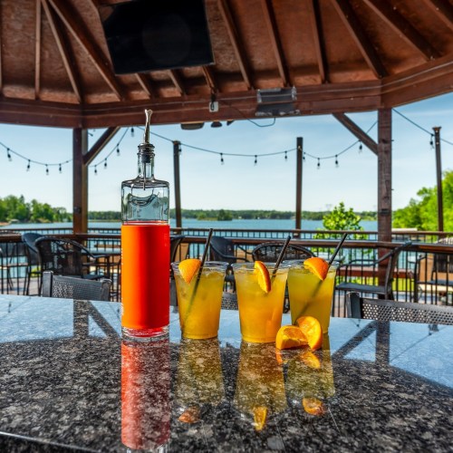 A bottle of red liquid next to three orange cocktails with slices, set on a table overlooking a lake.