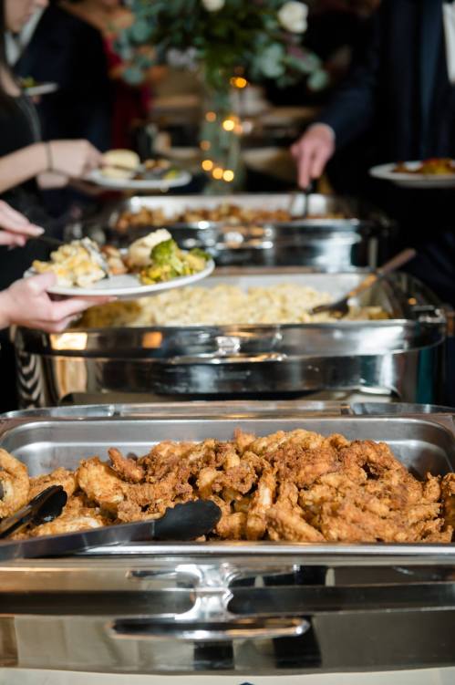 A buffet table with trays of fried chicken, pasta, and broccoli, as guests serve themselves.