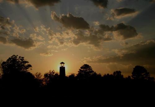 Silhouette of a tower against a sunset, with rays of light breaking through clouds and trees in the foreground.