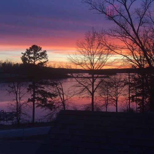 A serene lake at sunset, with vibrant pink and purple skies and silhouetted trees along the shoreline.