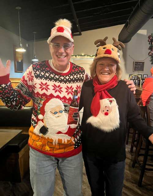 A man in a festive Santa sweater and a woman in a reindeer hat and holiday shirt smile and wave in a cheerful setting.