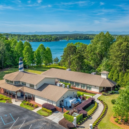 Aerial view of a lakeside building with a lighthouse, surrounded by greenery and water under a clear blue sky.