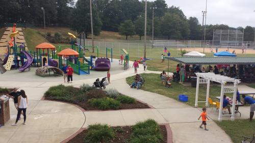 A vibrant park scene with children playing on colorful equipment, families enjoying the outdoors, and sports fields in the background.