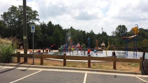Children playing in a splash pad at a park, with colorful water features and trees in the background.