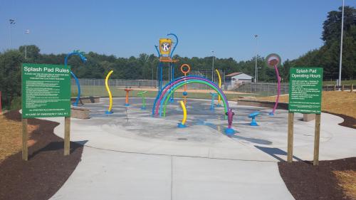 Colorful splash pad with water features, surrounded by green signs detailing rules and operating hours.