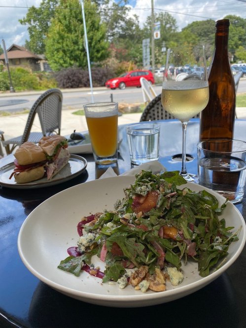 A plate of salad with greens, nuts, and cheese, alongside a sandwich, drinks, and a bottle on a table.