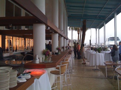 A waterfront restaurant with a bar, tables, and palm trees, featuring guests mingling and enjoying the atmosphere.