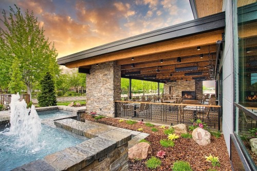 A modern outdoor patio with a stone fountain, lush greenery, and a cozy fireplace under a colorful sunset sky.