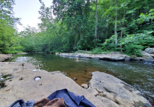 A serene river scene surrounded by lush greenery, with a person swimming in the water and large rocks nearby.