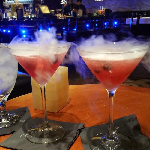 Two pink cocktails with smoke effect, served in martini glasses on a wooden table, with a bar in the background.