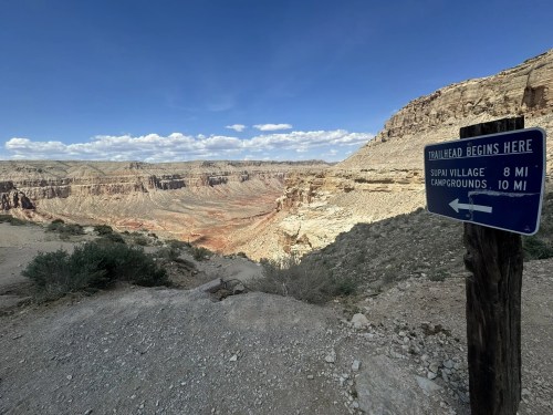 Trailhead sign pointing left, indicating Supai Village 8 miles and campgrounds 10 miles, with a canyon view in the background.