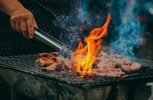 A hand holds tongs over a grill with sizzling meat and vibrant flames, surrounded by smoke.