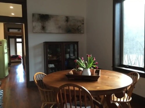 A cozy dining area featuring a round wooden table, chairs, and a vase of tulips, with a cabinet and artwork in the background.