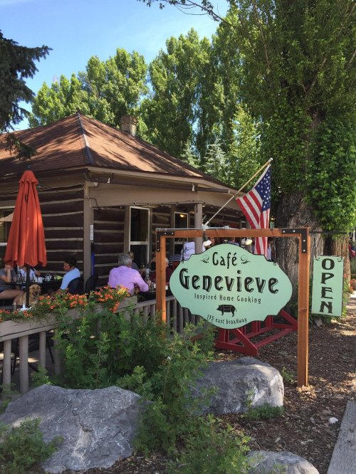 A rustic café sign reading "Café Genevieve" with an American flag, surrounded by greenery and outdoor seating.